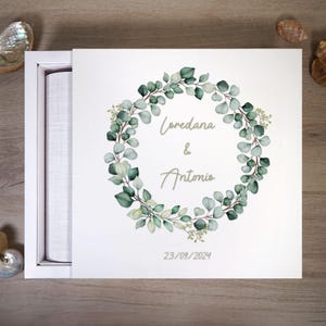 Può includere: Scatola bianca con coperchio bianco. Il coperchio è aperto e l'interno della scatola è rivestito di tessuto bianco. La parte anteriore della scatola presenta un'illustrazione ad acquerello di una ghirlanda di eucalipto verde con il testo "Loredana & Antonio" e la data "23/09/2024".