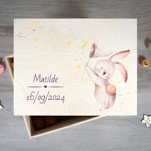 Peut inclure: Une boîte en bois avec une finition en bois clair. Le couvercle présente une illustration aquarelle d'un lapin tenant un filet à papillons. L'arrière-plan est jaune clair avec des étoiles dorées. Le texte "Matilde 16/09/2024" est imprimé sur le couvercle.
