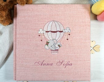 May include: A pink photo album with an embroidered hot air balloon design featuring an elephant and a bunny. The album cover has the name "Anna Sofia" embroidered in a cursive font. The words "It's a Girl" are on a wooden star.