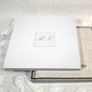 May include: White photo album box with a linen-textured cover. The box has a gold embossed inscription that reads "M.S."