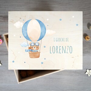 Peut inclure: Une boîte à jouets en bois avec une finition en bois clair. Le couvercle présente une montgolfière bleue et blanche avec un ours brun portant des lunettes d'aviateur et une écharpe bleue. Le texte "I GIOCHI DI LORENZO" est imprimé sur le couvercle.