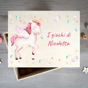 Peut inclure: Une boîte en bois avec une illustration de licorne et le texte "I giochi di Nicoletta" sur le couvercle. La licorne est rose avec une crinière et des ailes arc-en-ciel. La boîte est décorée de petits cœurs, d'étoiles et de pierres précieuses.
