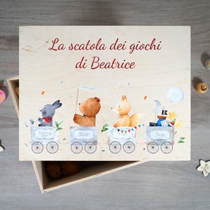 Peut inclure: Boîte à jouets en bois avec un design fantaisiste représentant un train avec des passagers animaux. La boîte est étiquetée "La scatola dei giochi di Beatrice".