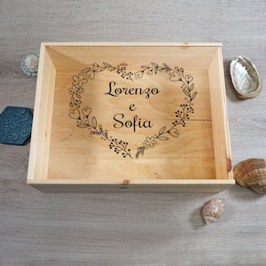 Op de afbeelding: Een houten doos met een hartvormig ontwerp gegraveerd met de namen "Lorenzo e Sofia" omringd door een krans van bloemen.