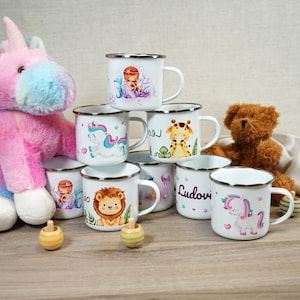 Könnte beinhalten: Ein Stapel weißer Emailletassen mit bunten Comic-Tiermotiven, darunter ein Einhorn, ein Löwe, eine Giraffe und eine Meerjungfrau. Die Tassen haben silberne Ränder und Griffe. Auf dem Tisch liegen zwei Holz-Kreisel.