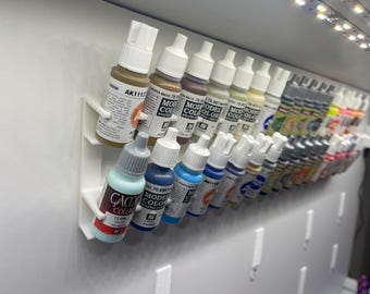 May include: A collection of acrylic paint bottles in various colors, including brown, blue, and beige, are displayed in a white organizer. The bottles are labeled with brand names and color codes. The image shows a variety of paint colors for model painting.