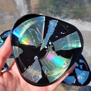 May include: A set of four black coasters made from broken CDs. The coasters have a iridescent, rainbow-like effect.