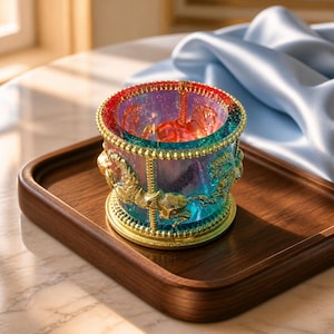 May include: A cylindrical candle holder featuring a carousel design with gold-toned horses and beaded trim. The glass body displays a gradient of iridescent blue, purple, and red hues with a subtle glitter finish throughout the structure.