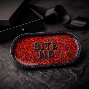 May include: An oval-shaped resin tray featuring a vibrant red glitter base with a dark, speckled border. The words BITE ME are embossed in the center in a bold, black, gothic-style font. The tray has a smooth, glossy finish and a raised edge.