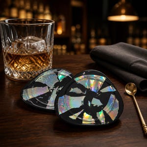 May include: Two triangular coasters with rounded edges featuring a black base inlaid with iridescent, reflective shards that create a prismatic effect. The coasters have a smooth, glossy finish and are arranged on a dark wooden surface.