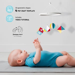 Può includere: Un bambino guarda un mobile sospeso con forme geometriche rosse, gialle, blu e bianche. L'immagine include il testo che recita "3D geometric shapes", "4 PDF CRAFT TEMPLATE" e "VIDEO TUTORIAL". Il bambino indossa un body blu.