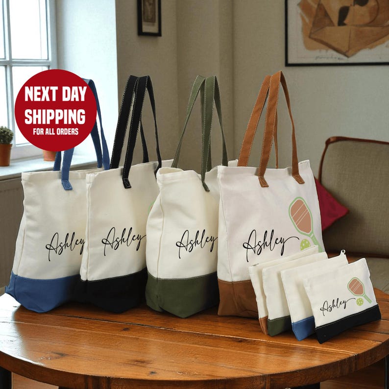 a collection of tote bags and a small bag, each with the name "Ashley" printed on them. The bags are arranged on a wooden table, with a sign indicating that they are available for shipping to all orders.