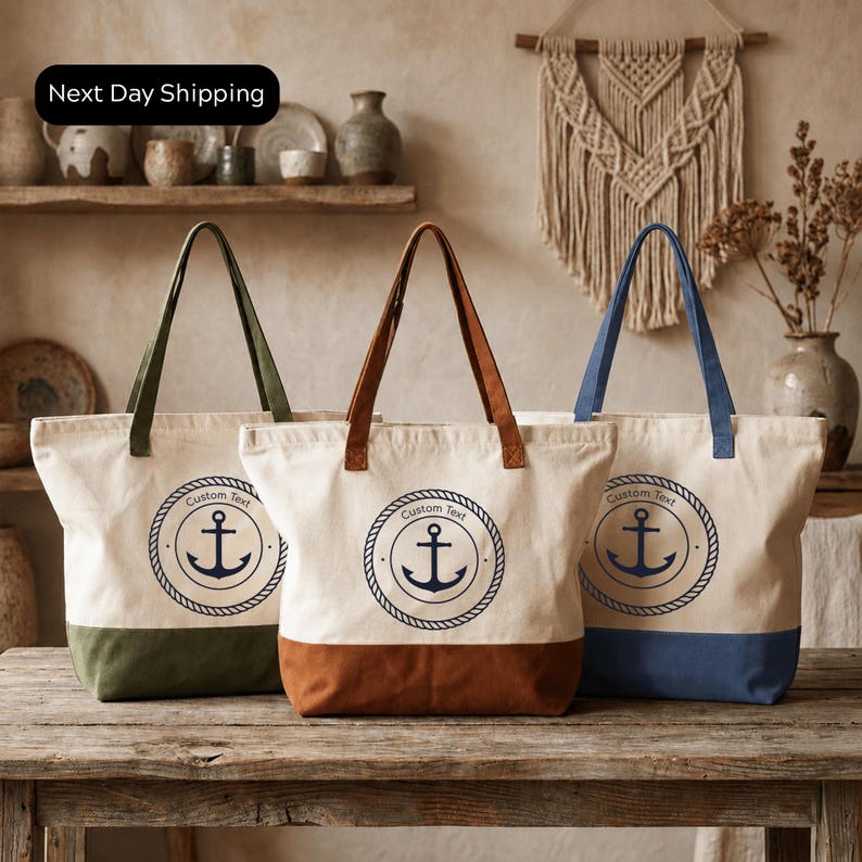 Three tote bags with a nautical design, featuring a blue anchor and a white anchor on a beige background. The bags are arranged on a wooden surface, with a shelf and a wall decoration in the background.
