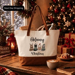 A white tote bag with a Christmas-themed design, featuring illustrations of cats and the text "Meowvy Christmas" on it, placed on a wooden table. The table also has a cup of coffee, a pastry, and a book. In the background, there is a Christmas tree with ornaments and lights, and a sign that says "Next Day Shipping".