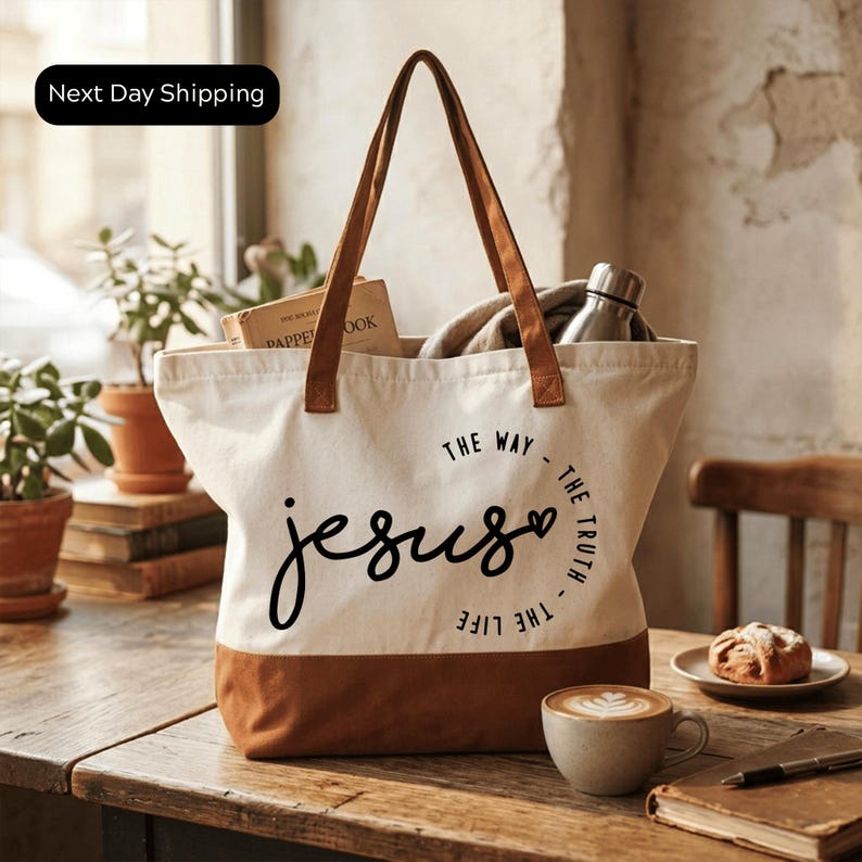 A white tote bag with the text "The way Jesus" printed on it, along with a coffee cup and a pastry on a wooden table.