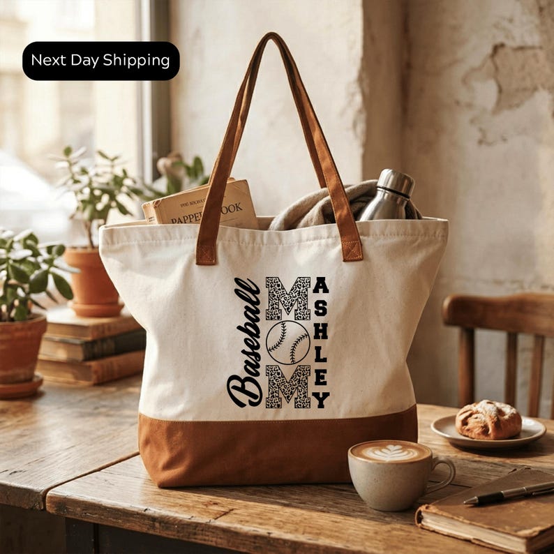 A white tote bag with the text "Baseball Mom Ashley" printed on it, sitting on a wooden table. The bag has a handle and appears to be filled with various items, including a book, a water bottle, and a pastry. There is also a cup of coffee and a plant in the background.