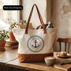 A white tote bag with a blue anchor symbol and the text "Custom Text" printed on it, placed on a wooden table. The bag is filled with various items, including a water bottle, a book, and a pastry. There is also a cup of coffee and a plant in the background.
