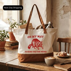 A white tote bag with a red raccoon graphic and the text "ON MY WAY TO MAKE THE VIBE WEIRD" printed on it, placed on a wooden table. There is a cup of coffee and a pastry nearby, and a potted plant in the background.