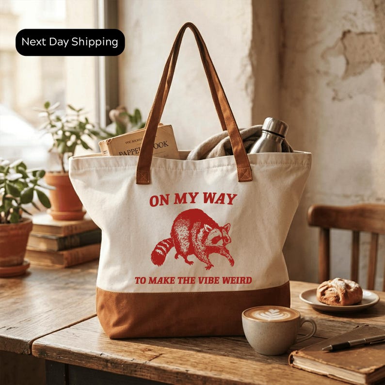 A white tote bag with a red raccoon graphic and the text "ON MY WAY TO MAKE THE VIBE WEIRD" printed on it, placed on a wooden table. There is a cup of coffee and a pastry nearby, and a potted plant in the background.