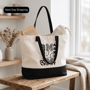 A beige tote bag with a black handle and straps, featuring a baseball design and the text "MAYBE" printed on it, is placed on a wooden surface.