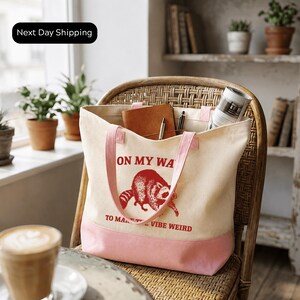 A white tote bag with a red raccoon design and the text "ON MY WAY TO MAKE IT VIBE WEIRD" is sitting on a wicker chair in a room with potted plants and a window.