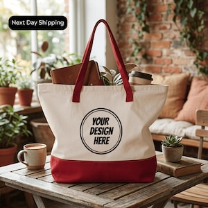 A white tote bag with a red trim, featuring the text "YOUR DESIGN HERE" printed on it, is placed on a wooden table. The bag is filled with various items, including a coffee cup, a book, and some plants. The setting appears to be a cozy indoor space with a brick wall and a window in the background.