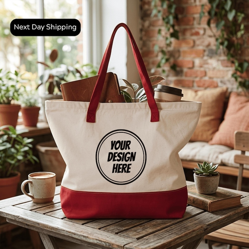 A white tote bag with a red trim, featuring the text "YOUR DESIGN HERE" printed on it, is placed on a wooden table. The bag is filled with various items, including a coffee cup, a book, and some plants. The setting appears to be a cozy indoor space with a brick wall and a window in the background.