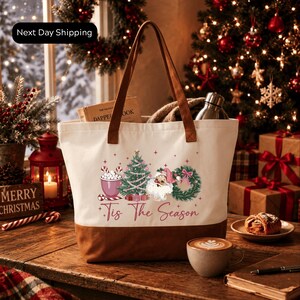 A white tote bag with a festive design, including a Christmas tree, Santa Claus, and the text "Tis The Season", placed on a wooden table. The table also has a cup of coffee, a pastry, and a book. The background features a decorated Christmas tree and other holiday decorations.