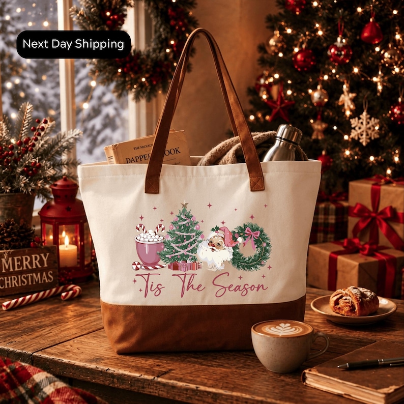 A white tote bag with a festive design, including a Christmas tree, Santa Claus, and the text "Tis The Season", placed on a wooden table. The table also has a cup of coffee, a pastry, and a book. The background features a decorated Christmas tree and other holiday decorations.