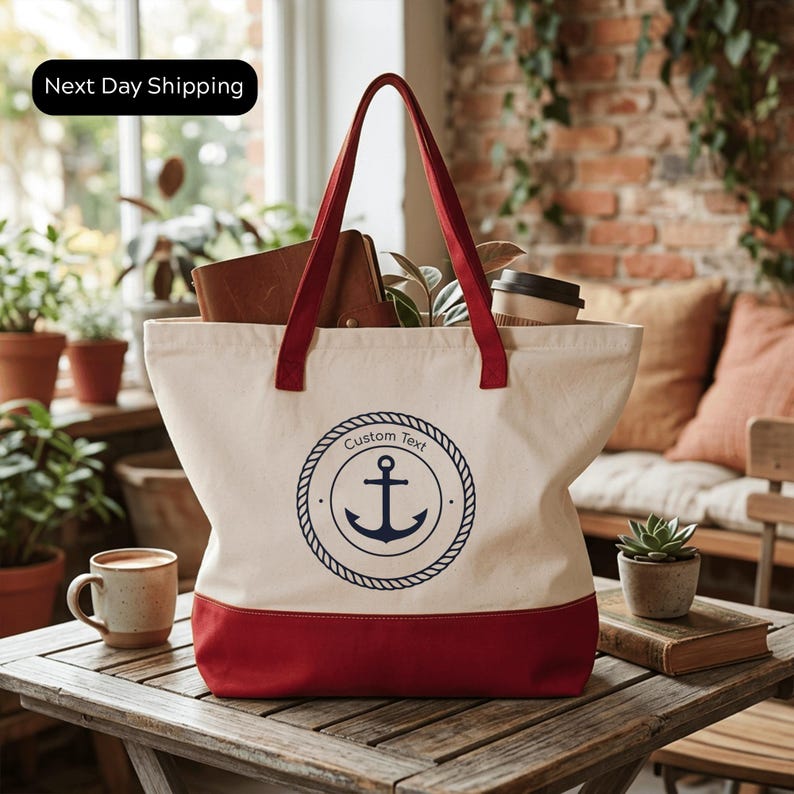 A red and white tote bag with a navy blue anchor symbol sits on a wooden table, surrounded by potted plants and a coffee cup.