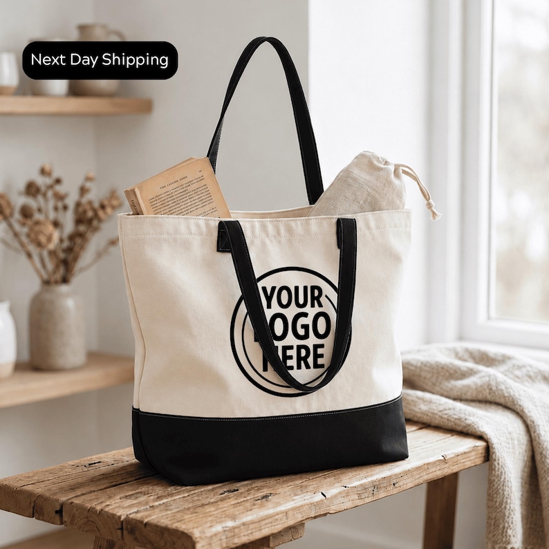 A beige tote bag with a black handle and trim, featuring a logo on the front, is placed on a wooden surface.