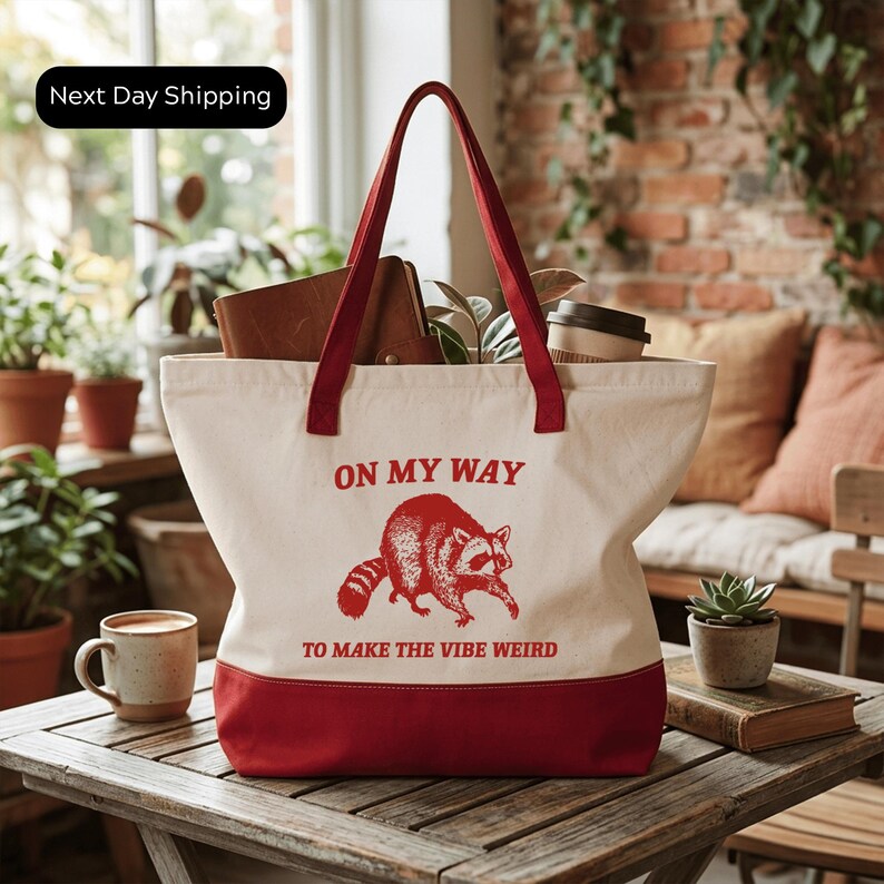 A red and white tote bag with the text "ON MY WAY TO MAKE THE VIBE WEIRD" printed on it, placed on a wooden table in a room with potted plants and a brick wall.