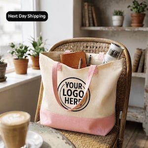 A beige tote bag with a pink trim, featuring a logo, is placed on a wicker chair in a room with potted plants and a window.