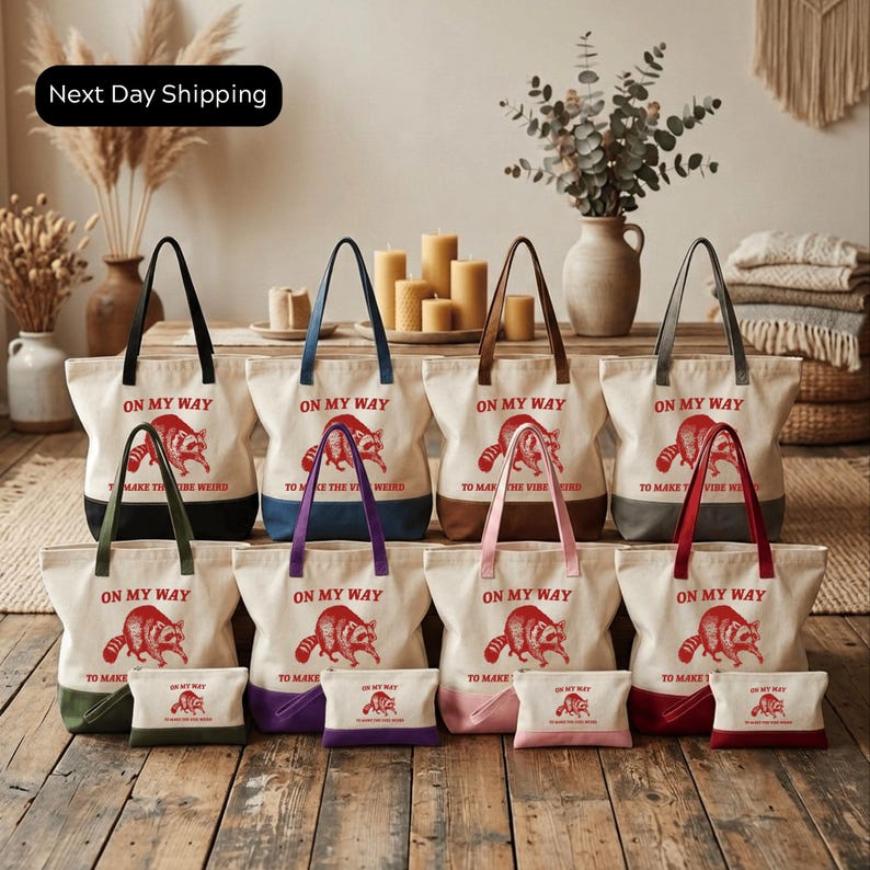 A collection of white tote bags with red text and images of raccoons, arranged on a wooden floor in front of a vase with greenery and candles.