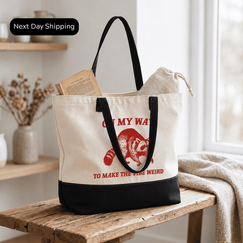 A white tote bag with a red raccoon graphic and the text "ON MY WAY TO MAKE THE VIBE WEIRD" printed on it, placed on a wooden surface.