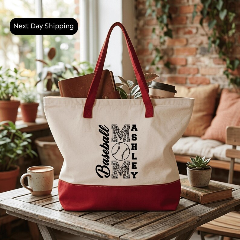 A red and white tote bag with the text "Baseball Mom" printed on it, sitting on a wooden table in a room with potted plants and a brick wall.