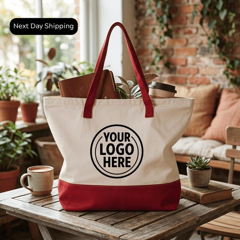 A white tote bag with a red trim, featuring a black circular logo that says "YOUR LOGO HERE". The bag is placed on a wooden table, with a coffee cup and a plant nearby.