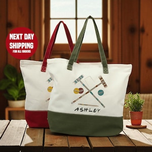 May include: Two canvas tote bags with white bodies, red and green handles, and color-blocked bottoms. Each bag features a croquet mallet and ball design, with the name "ASHLEY" printed on one. The bags are on a wooden table.