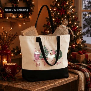 A white tote bag with a Christmas-themed design, including a mug with a hot beverage, a Christmas tree, and a Santa Claus figure, is placed on a wooden surface.