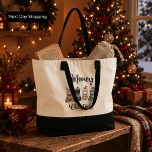 A white tote bag with a black handle and a printed design of cats is placed on a wooden surface, with a Christmas tree and holiday decorations visible in the background.