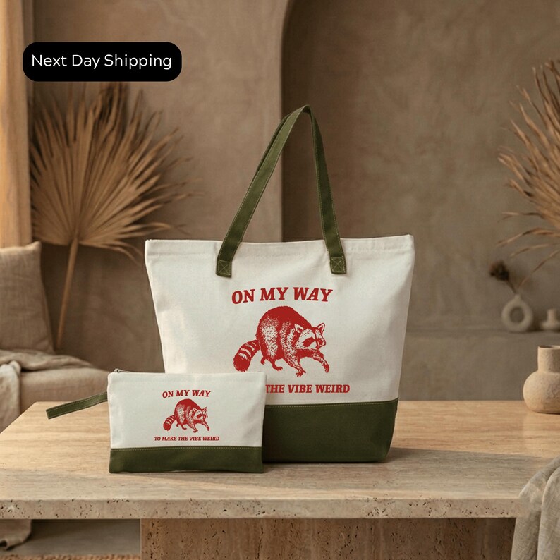 A white tote bag with a green handle and a smaller white bag with a red design featuring a raccoon and the text "ON MY WAY" and "THE VIBE WEIRD" printed on it, placed on a wooden table.