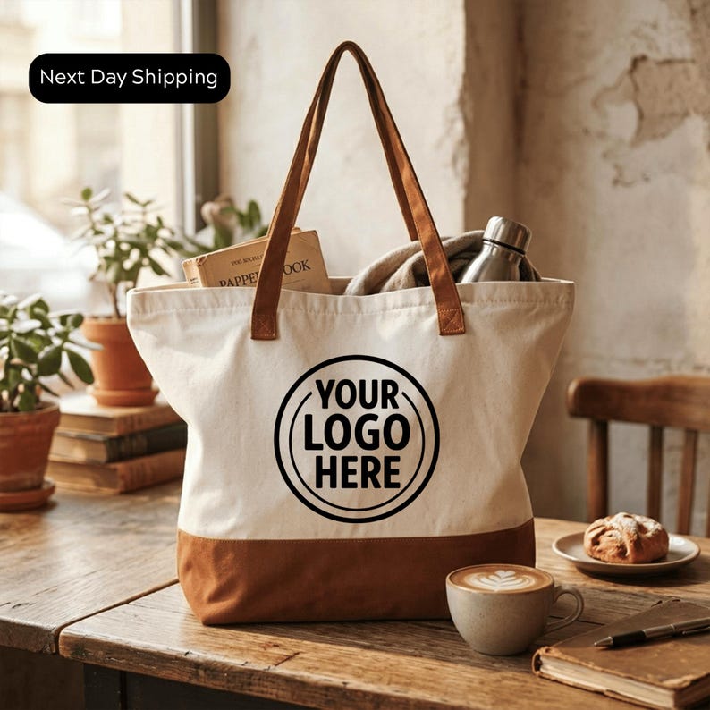 A white tote bag with the text "YOUR LOOP HERE" printed on it, placed on a wooden table. The bag is filled with various items, including a coffee cup, a pastry, and some books. The table also has a plant and a stack of books.