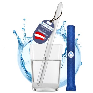 Pode incluir: Uma palhinha de vidro transparente é colocada num copo com água. Uma etiqueta azul com o texto "nomavital HANDARBEIT AUS ÖSTERREICH" e a bandeira austríaca pendura-se da palhinha. Um filtro azul está ao lado do copo. A água salpica à volta do copo.