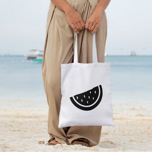 K&ouml;nnte beinhalten: Wei&szlig;e Tragetasche mit einem schwarzen Wassermelonen-Scheiben-Design. Die Tasche wird von einer Person an einem Sandstrand gehalten. Im Hintergrund sind das Meer und Boote zu sehen. Eine einfache, stilvolle Tragetasche.