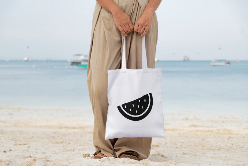 K&ouml;nnte beinhalten: Wei&szlig;e Tragetasche mit einem schwarzen Wassermelonen-Scheiben-Design. Die Tasche wird von einer Person an einem Sandstrand gehalten. Im Hintergrund sind das Meer und Boote zu sehen. Eine einfache, stilvolle Tragetasche.