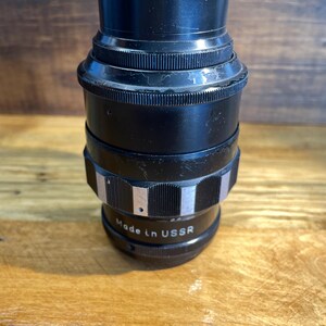 Op de afbeelding: Een zwarte cameralens met een geribbeld ontwerp en zilveren accenten. De lens heeft de tekst "Made in USSR" op de zijkant. De lens is cilindrisch en staat op een houten oppervlak.