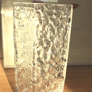 Può includere: Un vaso di vetro trasparente e rettangolare con una superficie strutturata simile al ghiaccio. Il vaso è appoggiato su una superficie di legno. Lo sfondo include un'opera d'arte incorniciata con una serie di labbra rosse.