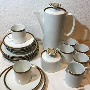 Op de afbeelding: Een witte keramische koffieset met bruine rand. De set bevat een koffiepot, suikerpot, melkkan, kopjes en schotels. De koffiepot en suikerpot hebben decoratieve deksels. De set staat op een wit tafelkleed.