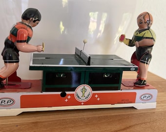 Peut inclure: Un jouet en fer-blanc de style vintage représentant deux personnages jouant au tennis de table sur une table verte avec un filet et une petite balle. La base est orange et blanche avec le texte P.P, un emblème de trophée et MS 334 MADE IN CHINA.