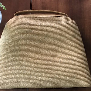 Könnte beinhalten: Eine hellbraune gewebte Handtasche mit gewölbtem Oberteil und Griff. Die Tasche hat eine abgerundete, trapezförmige Form und besteht aus dicht gewebtem Stroh. Der Hintergrund ist eine Holzoberfläche.